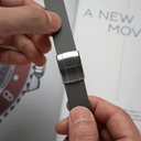 Close-up of a person's hands holding a dark gray watch strap with a brushed silver metal clasp, with a blurred magazine in the background.