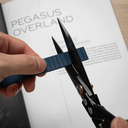 A close-up of a hand holding a dark blue textured strip while using black-handled shears to trim it, with a printed page in the background showing the text 'PEGASUS OVERLAND'.