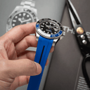 A wristwatch with a bright blue strap and black dial held by a hand. The watch has a silver-toned metal case with a black and blue rotating bezel.