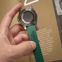 A hand holding a silver-toned watch case with a green strap, shown close-up against a brown paper background with printed text.