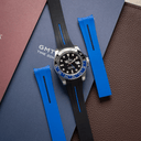 A wristwatch with a black dial, blue and black bezel, and black rubber strap with blue center lines, arranged on top of a blue GMT Time Zone booklet with two blue strap pieces, against a brown textured surface.